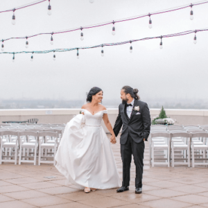 A newlywed couple holding hands and smiling at each other on a rooftop with string lights above and chairs arranged for their wedding in the background.