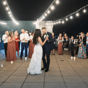 A couple shares their first dance under string lights as guests look on at an evening wedding reception.