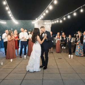 A newlywed couple shares their first dance at their wedding outdoors surrounded by guests under string lights.