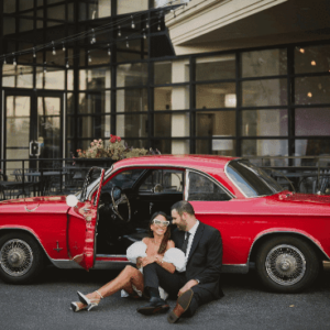 A couple sitting on the ground, leaning against a classic red car, sharing an affectionate moment.