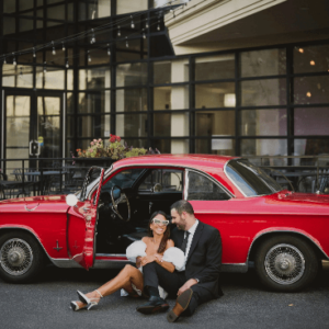 A newlywed couple sits beside a classic red car on an urban street.