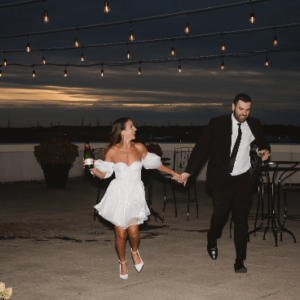 A couple holding hands and running at nighttime under string lights at their wedding, with the man carrying a bottle.