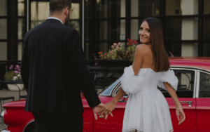 A bride in a white dress holding hands with a groom in a suit, looking back over her shoulder, with a classic red car and a building in the background.