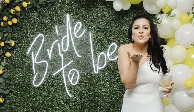 A bride-to-be blowing a kiss, standing in front of a decorative backdrop with the phrase "bride to be" and balloons at her wedding shower.