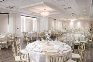 An elegantly set wedding event hall with round tables, gold chairs, and white decor.