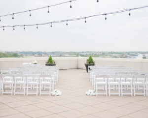 Outdoor rooftop wedding event space set up with rows of white chairs for a ceremony, strung lights above, and a city skyline in the distance.