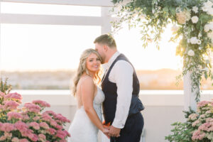 Bride and groom holding hands amidst floral arrangements in soft sunset light at their wedding.